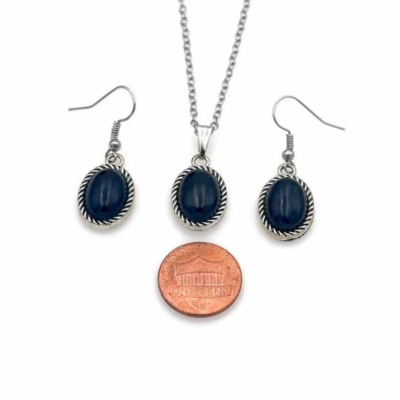 May include: A silver necklace and earrings set with black oval stones. The pendant and earrings are surrounded by a silver rope design. The set is shown next to a US penny for size comparison.