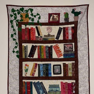 Puede incluir: Un tapiz de pared acolchado que representa una estantería llena de libros coloridos. Los estantes marrones contienen varios libros, fotos enmarcadas y objetos decorativos. El fondo es de color crema con escritura manuscrita.