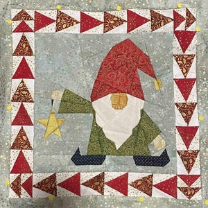 May include: A quilted wall hanging featuring a gnome holding a star. The gnome is wearing a red hat with gold accents, a green jacket, and black boots. The background is a light blue with a white snowflake pattern. The border is made of red and gold triangles.