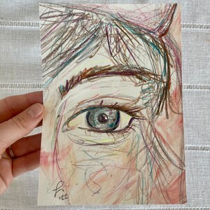 May include: A pastel drawing of an eye, with a focus on the iris and surrounding details. The artwork features a mix of colors, including pink, blue, and brown, creating a soft and expressive portrait. The artist's signature and the year '22 are visible.