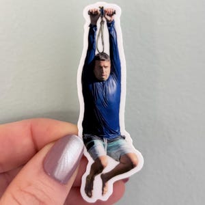Pode incluir: Um autocolante de vinil recortado que mostra uma pessoa com uma camisola azul-escura de manga comprida e calções de banho azul-claros, pendurada numa pega metálica com uma corda. A pessoa tem uma expressão neutra e pernas dobradas.