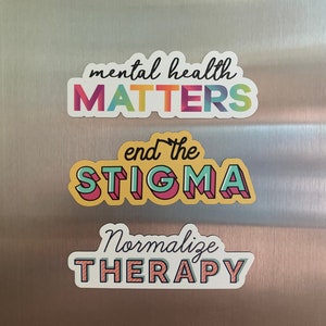 Può includere: Tre adesivi colorati con i testi "mental health MATTERS", "end the STIGMA" e "Normalize THERAPY". Gli adesivi hanno un bordo bianco e sono in vari colori, tra cui rosa e verde. Lo sfondo è una superficie in metallo spazzolato.