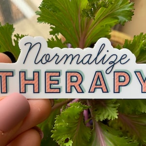 May include: A sticker with the words "Normalise THERAPY" in a stylized font. The word "Normalise" is in a cursive font, and the word "THERAPY" is in a block font. The sticker has a white background with a blue border and is held in front of green leafy plants.