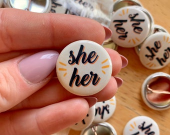 Può includere: Una collezione di bottoni rotondi bianchi con la scritta "she her" in corsivo nero, accentuata da raggi arancioni. I bottoni sono tenuti in mano, con altri visibili sullo sfondo.