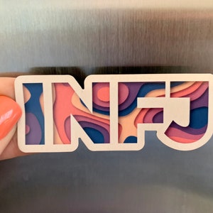 May include: A white magnet with the letters "INFJ" in a layered paper design. The layers are in shades of pink, purple, and blue. The magnet is held against a metallic surface.