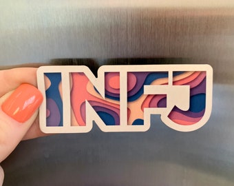 Puede incluir: Un imán blanco con las letras "INFJ" en un diseño de papel en capas. Las capas son en tonos de rosa, morado y azul. El imán se sostiene contra una superficie metálica.