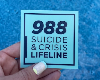 May include: A square sticker with a light teal background and a black border. The number "988" is prominently displayed at the top, followed by the words "SUICIDE & CRISIS LIFELINE" in a stacked format. The sticker is held by a hand.