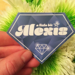Op de afbeelding: Een blauw-witte diamantvormige sticker met de tekst "a little bit Alexis" in een retro lettertype. De sticker heeft een geometrisch patroon en een diamantafbeelding. De sticker wordt voor een groen-witte bloem gehouden.