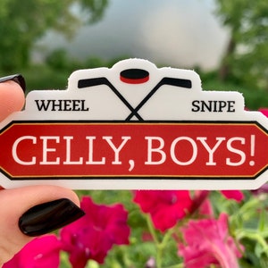 May include: A white sticker with a red and black design. The sticker features crossed hockey sticks, a hockey puck, and the words "WHEEL SNIPE" above a red rectangle with the words "CELLY, BOYS!" in white.