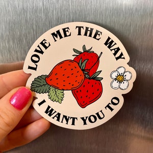 Peut inclure: Un autocollant rond avec le texte "LOVE ME THE WAY I WANT YOU TO" entourant un groupe de trois fraises rouges, des feuilles vertes et une fleur blanche avec un centre jaune.