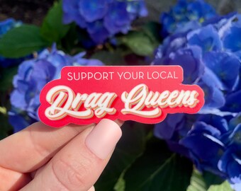 Pode incluir: Um autocolante vermelho com o texto "SUPPORT YOUR LOCAL Drag Queens" em letras brancas e amarelas. O autocolante é segurado em frente a um fundo de flores azuis.