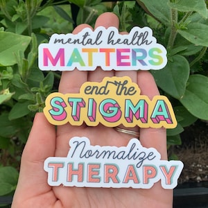 Könnte beinhalten: Drei farbenfrohe Aufkleber mit Texten zur Förderung des Bewusstseins für psychische Gesundheit. Der obere Aufkleber lautet "mental health MATTERS" in Regenbogenfarben. Der mittlere Aufkleber lautet "end the STIGMA" und der untere Aufkleber lautet "Normalize THERAPY".