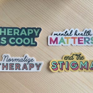 May include: Four stickers with mental health messages. The stickers read: "THERAPY IS COOL", "mental health MATTERS", "Normalise THERAPY", and "end the STIGMA". The stickers have a white border and are on a light wood surface.