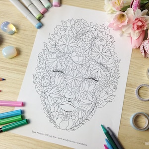 K&ouml;nnte beinhalten: Eine Schwarzwei&szlig;-Zeichnung eines Gesichts mit Blumen und Bl&auml;ttern, die das gesamte Gesicht bedecken. Die Zeichnung befindet sich auf einem wei&szlig;en Blatt Papier. Das Papier ist von Kunstmaterialien umgeben, darunter farbige Bleistifte, Marker und Washi-Tape.