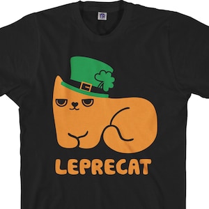 Puede incluir: Camiseta negra con un gato naranja que lleva un sombrero verde de duende con un trébol. El texto "LEPRECAT" está impreso debajo del gato.