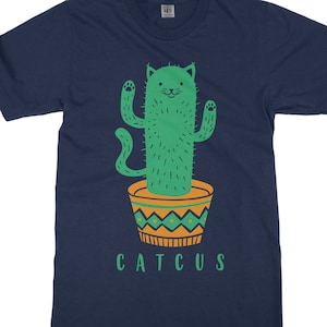 May include: A navy blue t-shirt featuring a graphic of a green cat-shaped cactus in a yellow and green pot. The text "CATTUS" is printed below the image.