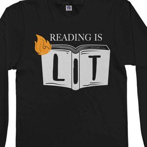以下が含まれることがあります： 炎と「Reading is Lit」という文字が書かれた本の白いグラフィックがプリントされた、黒い長袖Tシャツです。