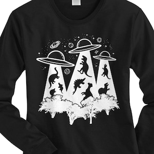 Puede incluir: Camiseta negra de manga larga con un gráfico blanco de tres ovnis flotando sobre un grupo de dinosaurios en un bosque. Los ovnis tienen un diseño de estrella en la parte superior.