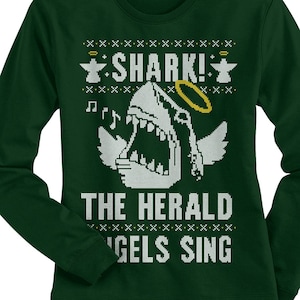 May include: Green long-sleeved shirt with a white shark wearing a halo and singing. The text "SHARK! THE HERALD 'GELS SING" is printed on the shirt.