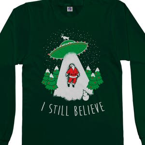 May include: A green long-sleeved t-shirt with a graphic of Santa Claus being abducted by a UFO. The graphic also includes a reindeer and Christmas trees. The text "I Still Believe" is printed below the graphic.