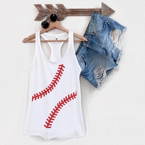 May include: White tank top with a red baseball stitch design. The tank top is hanging on a wooden arrow with a pair of sunglasses.