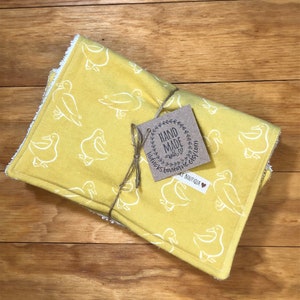 May include: A yellow fabric baby burp cloth with a white duck print. The cloth is folded and tied with twine. A brown paper tag is attached to the twine with the text "Hand Made with Love" and "Little Ducks Boutique".