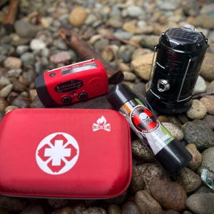 May include: A collection of emergency preparedness items, including a red first aid kit with a white cross, a red hand-crank radio, a black solar-powered lantern, and a black spray canister with a skull logo. The items are arranged on a bed of rocks.