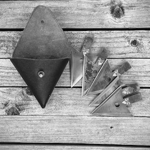 May include: A black leather pouch with a triangular flap and a snap closure. The pouch is lying on a wooden surface with a set of six metal throwing knives arranged in a fan shape.