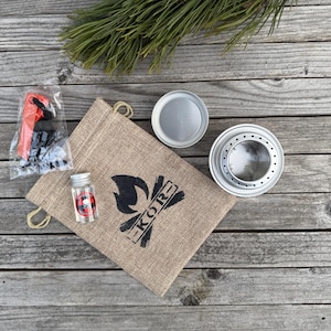 Peut inclure: Un ensemble de matériel de camping dans un sac en toile de jute brun avec le mot "KORE" imprimé dessus. L'ensemble comprend un petit réchaud en métal, une tasse en métal, une petite bouteille en verre et un allume-feu rouge et noir.