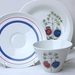 May include: A white ceramic teacup, saucer, and plate set. The plate has blue and red concentric circles. The cup and plate feature a whimsical design of red and blue bulbous shapes with green floral accents. A classic tableware set.
