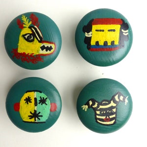 May include: Four round wooden knobs with different painted designs. Each knob features a different colourful face with geometric patterns and details. The knobs are teal green with yellow, red, black, and white accents.