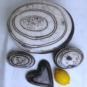 Puede incluir: Un juego de cuatro platos de cerámica en un esquema de colores blanco y negro. Los platos tienen forma de corazón, óvalo y dos óvalos más pequeños. Los platos tienen un esmalte craquelado y un borde negro. Un limón amarillo se encuentra en la mesa junto a los platos.