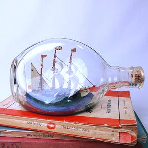 May include: A clear glass bottle containing a miniature sailboat with three masts and flags. The bottle has a cork stopper and sits atop a stack of vintage books with red and white covers. The ship is set on a blue base.