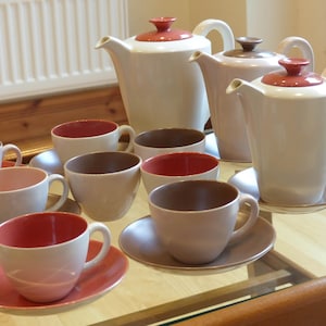 May include: A set of six teacups and saucers in various shades of brown, pink, and white. The cups and saucers have a smooth, rounded design. There are also three teapots in the background, two in white and one in pink.