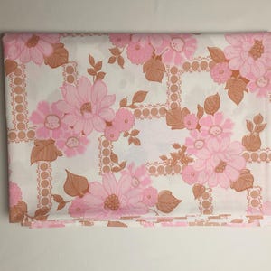 Op de afbeelding: Gevouwen bloemenstof met een herhalend patroon van grote roze bloemen en bruine bladeren op een witte achtergrond. Het ontwerp bevat een raster van bruine stippellijnen. Geschikt voor beddengoed of woondecoratie.