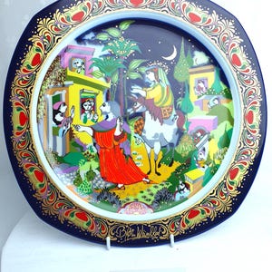 Op de afbeelding: Decoratieve plaat met een donkerblauwe rand en een kleurrijke illustratie. De plaat toont een scène met figuren, gebouwen en bomen, met gouden en rode accenten. Het jaar "1989" staat onderaan gedrukt.