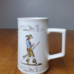 Könnte beinhalten: Eine weiße Keramiktasse mit Henkel. Die Tasse hat eine Schwarzweißillustration eines Mannes mit Hut, der eine Waffe hält. Die Illustration ist umgeben von anderen Schwarzweißillustrationen von Hüten, einem Bierkrug und einem Schwert.