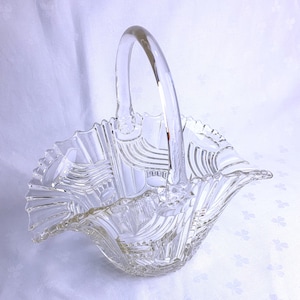 May include: Clear glass basket with a curved handle. The basket has a ruffled edge and a textured, geometric design. The handle is arched and attached to the basket's sides. The basket is likely for decorative use or for holding small items.