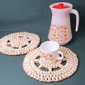 Puede incluir: Una jarra blanca con tapa roja y una taza blanca, ambas decoradas con motivos florales naranjas y rosas, sobre una superficie verde oscuro. También hay dos posavasos redondos hechos de semillas de color claro.