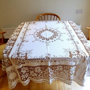 Puede incluir: Mantel rectangular blanco con diseño de encaje floral marrón. El encaje presenta un medallón central y un borde floral. El mantel cubre una mesa de madera. Adecuado para cenas u ocasiones especiales.