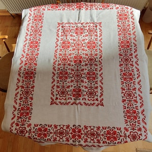 May include: A white tablecloth with a red floral pattern. The tablecloth is rectangular and covers a round table. The floral design is repeated around the edges and in the center of the tablecloth. The tablecloth is made of a woven fabric.