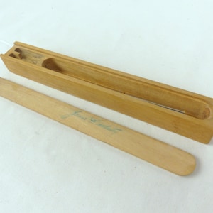 May include: A wooden box with a lid and a small wooden stick inside. The box is labeled "Jane S. Roberts".