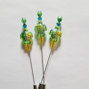 May include: Three decorative hat pins featuring iridescent green turtle charms. Each pin has a silver needle, a yellow accent, and is topped with a green faceted bead and blue crystal beads. The pins are displayed on a white background.