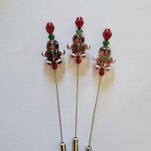 May include: Three Christmas-themed hat pins. Each pin features a gingerbread man charm with red and green accents, topped with a red bead and green crystal. The pins have silver-colored metal shafts and cylindrical bases.