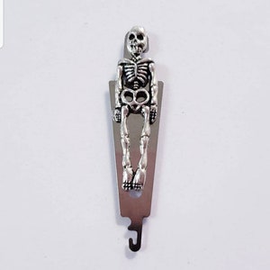 May include: A silver metal skeleton-shaped threader for sewing projects. The threader has a hook on the bottom and a skeleton design on the top.