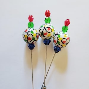 May include: Three decorative hat pins with white, patterned beads featuring red, yellow, and green accents. Each pin has a blue crystal base and is topped with a red and green bead cluster. The pins have silver-colored shafts.