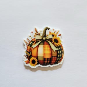 May include: A decorative pumpkin sticker with a plaid pattern in orange, yellow, and green. The pumpkin is adorned with a beige bow, sunflowers, and fall foliage. The sticker is on a white background.