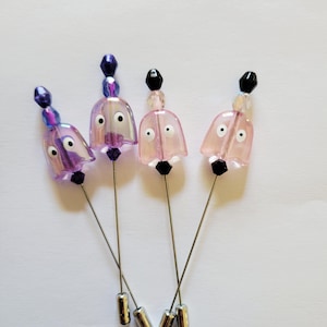 May include: Four decorative hat pins with bell-shaped heads. The heads are translucent, with a pink or purple hue, and feature small, round eyes. Each pin has a beaded top and a silver-colored pin.