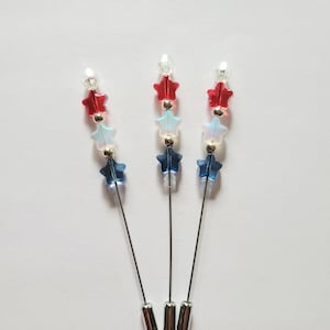 May include: Three decorative pins with star-shaped beads in red, white, and blue. Each pin has a silver-colored head and a long, thin, silver-colored pin. The star beads are arranged in a patriotic color scheme.