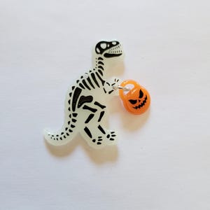 May include: A white glow-in-the-dark dinosaur skeleton holding a small orange pumpkin with a jack-o'-lantern face. The dinosaur is standing on a white background.